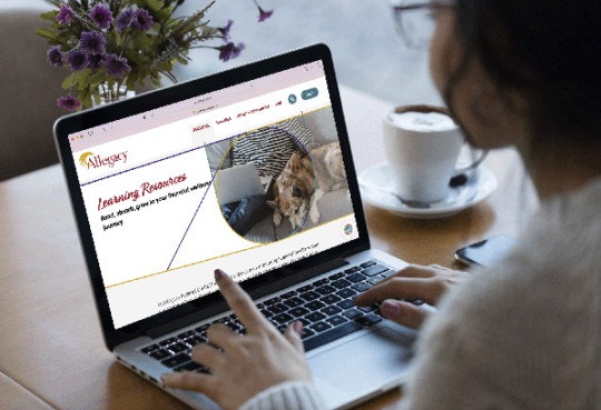 side profile of a woman wearing glasses and looking at laptop screen showing Allegacy's website with flowers and a coffee nearby on the table