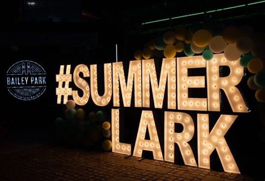 close up picture of the letters #Summerlark in theater lights near a balloon arch and the lighted Bailey Park sign