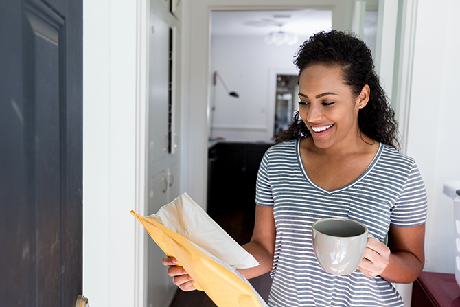 mid adult woman carrying a cup of coffee in sunny hallway smiling while looking at mail