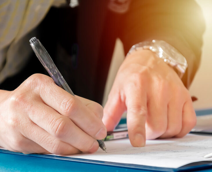 close up of a man holding the place on document with pointer finger of one hand while the other hand signs with a pen