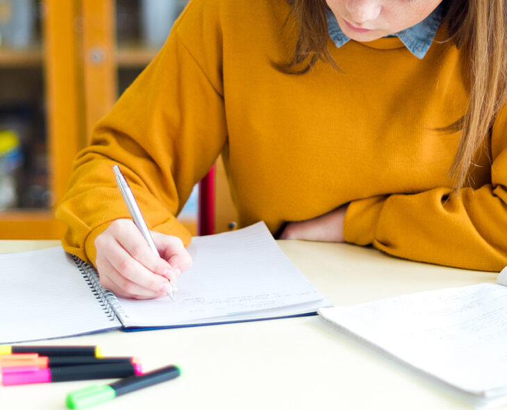 close up of student in gold sweatshirt writing in notebook with highlighters on table in foreground