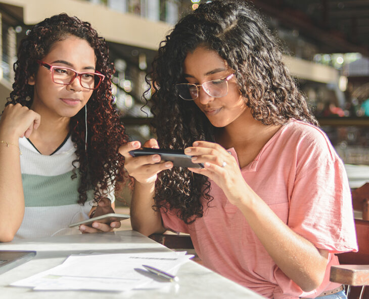 two young adults sit at a cafe table one taking a picture of a document with her phone while the other looks on