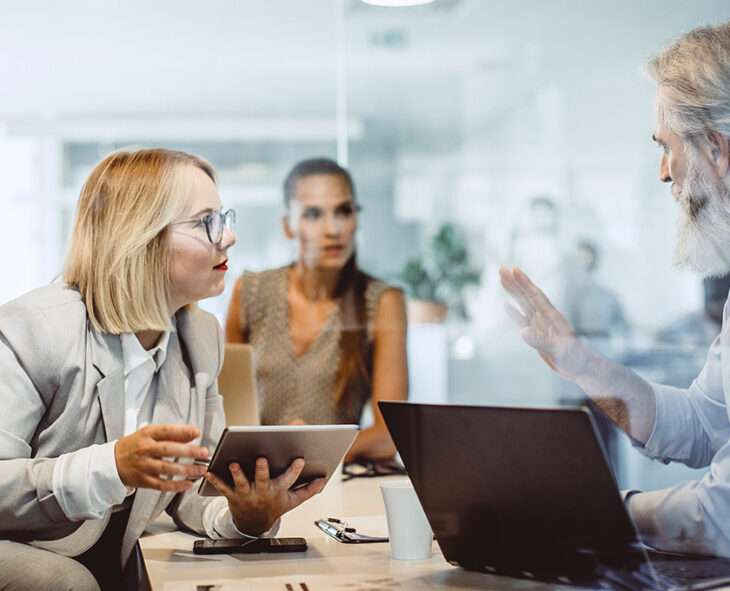 bespectacled woman holding tablet leaning on table with mobile phone and papers speaking with man using laptop across the table another employee sits in the background