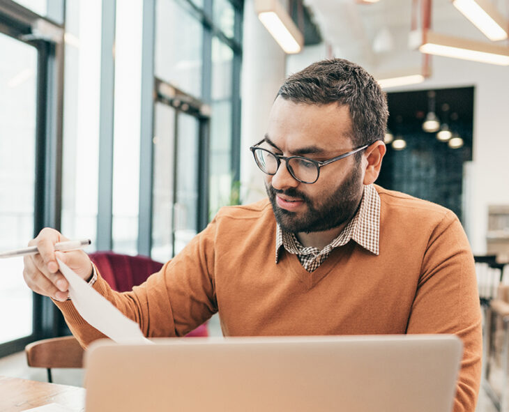 bespectacled man in front of laptop in large airy and bright office space looking at a piece of paper