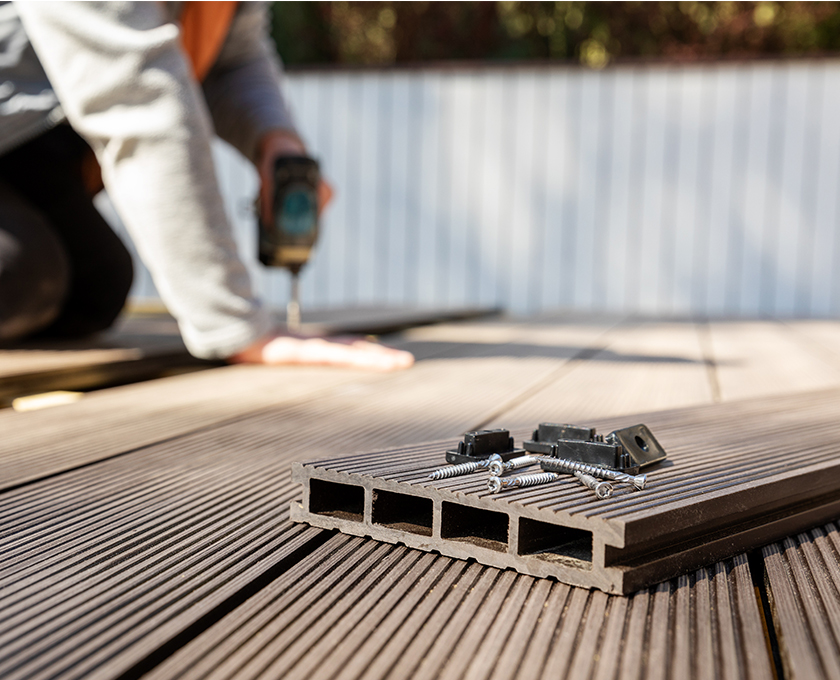 close up of weatherproof decking and screws in foreground while man holds board in place and drills in out of focus background