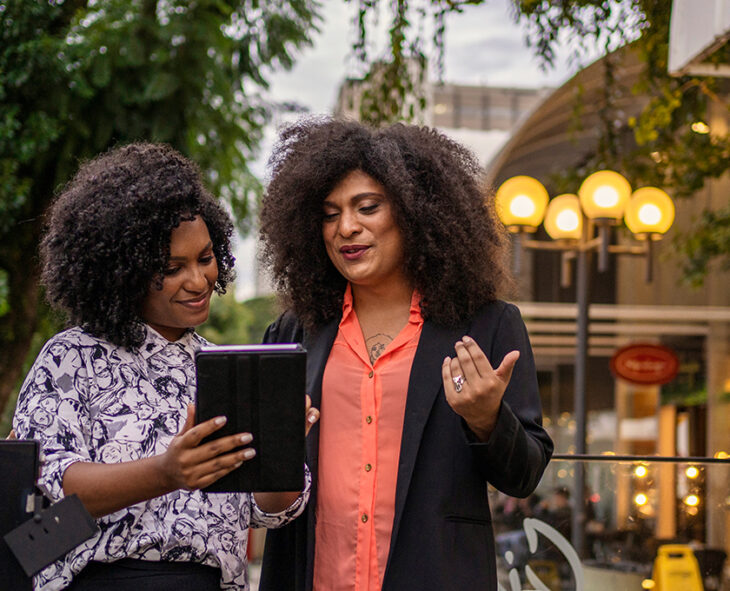 two business women standing outdoors near courtyard discussing something on tablet they are looking at