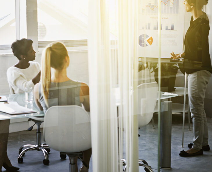 looking through an office window into a conference room where one woman is presenting a report to two other employees seated and looking on