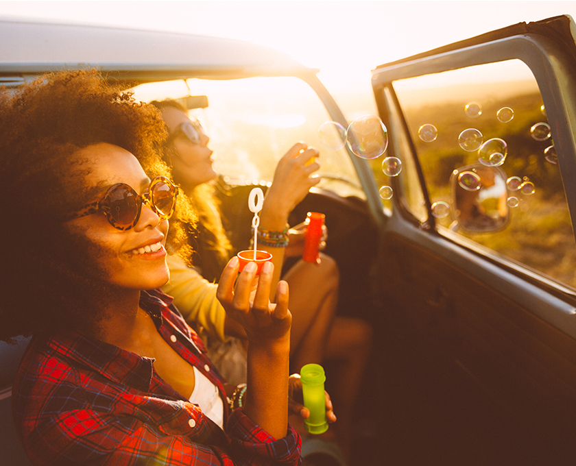 two young friends in parked jeep at sunset smiling and blowing bubbles into the wind
