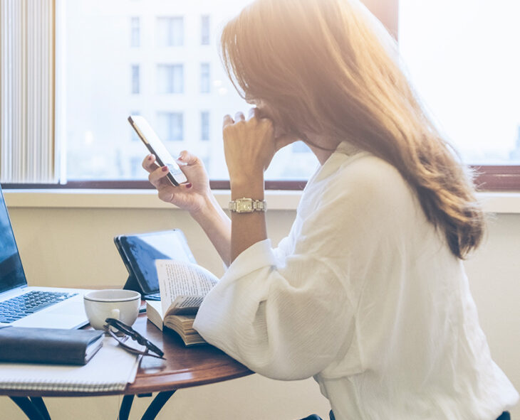 side profile of seated woman in office building checking her phone with laptop, glasses and coffee on the table in front of her