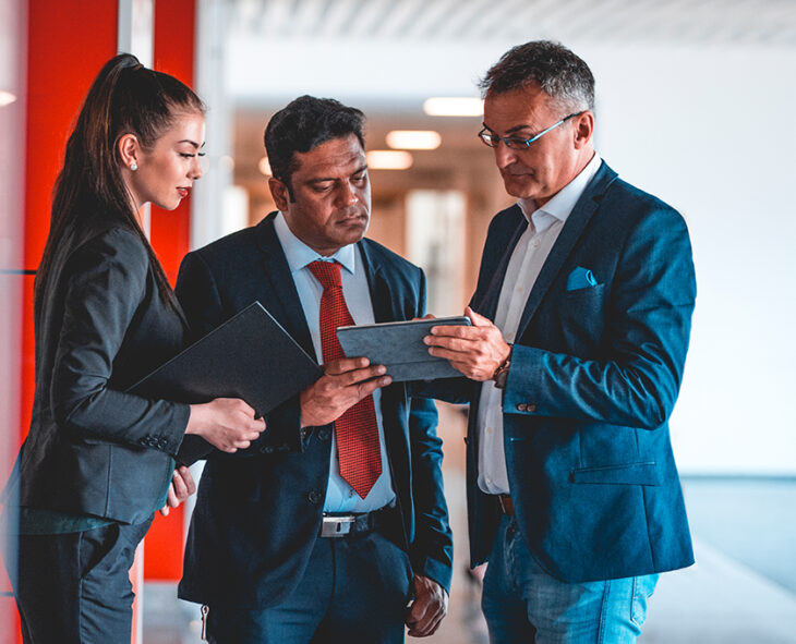 three employees in blazers stopped in red hallway looking at one man's tablet