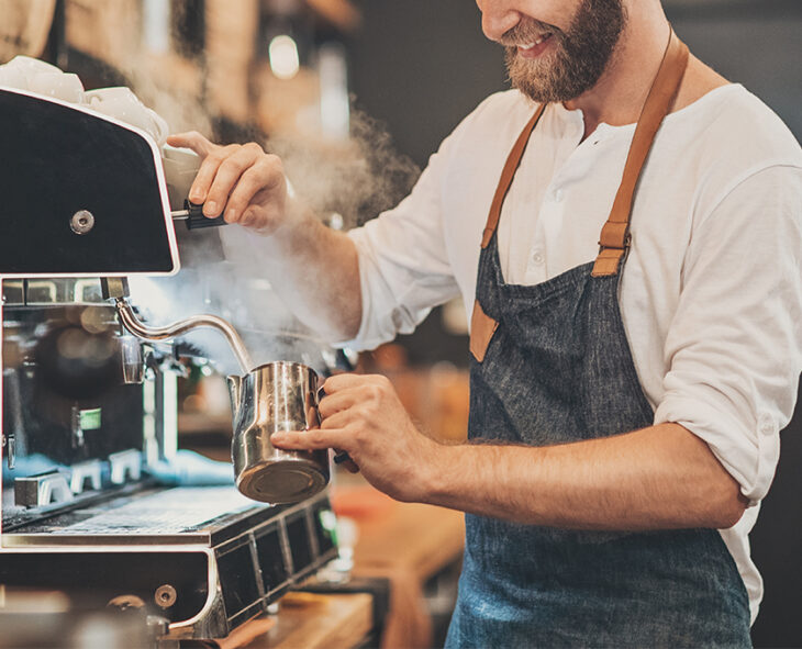 smiling bearded man with apron at coffee shop steaming milk