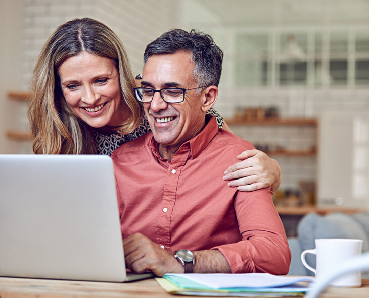 husband sits at kitchen table smiling at open laptop while wife leans over him with arm around his shoulder