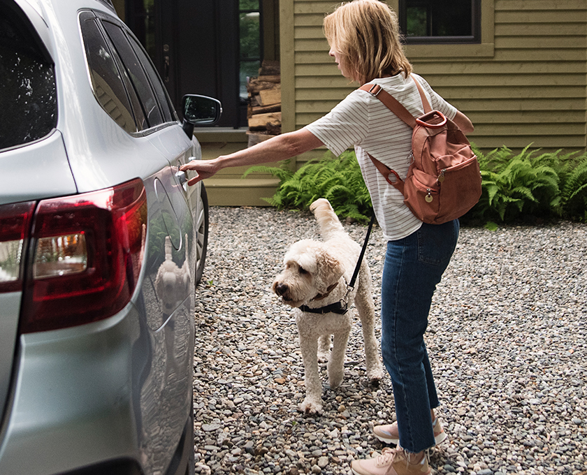 woman with backpack and sneakers on reaching for car door outside her home dog on lead waits to get in