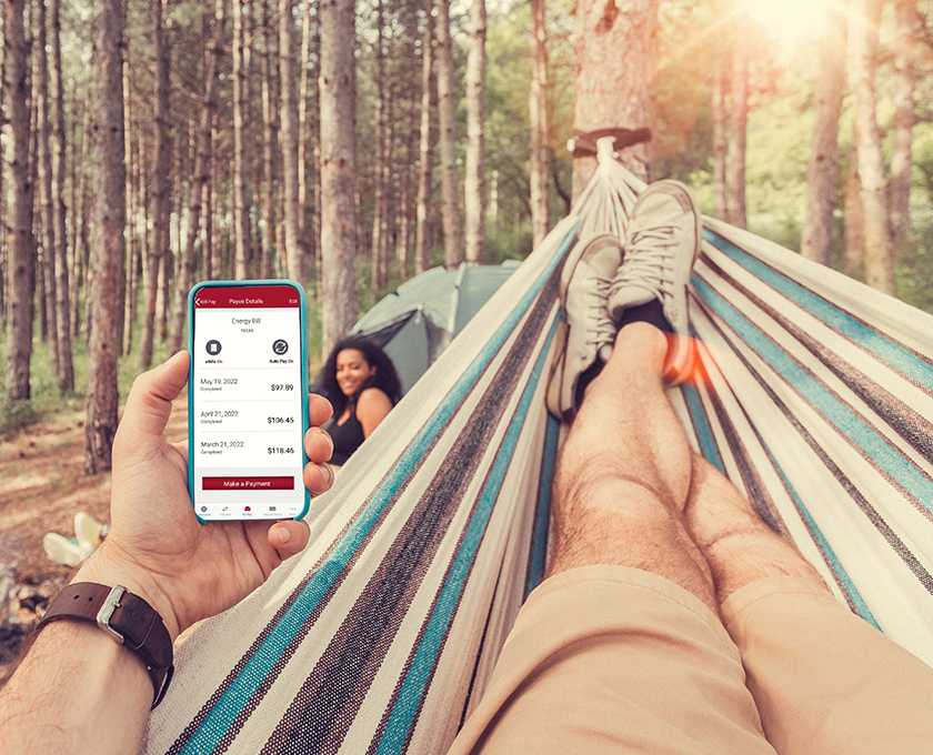 man laying in hammock in the woods looking at bill payment on a mobile phone with female in background by a tent