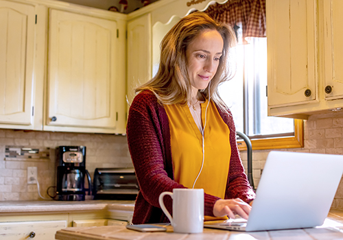 Woman in bright kitchen working on laptop.