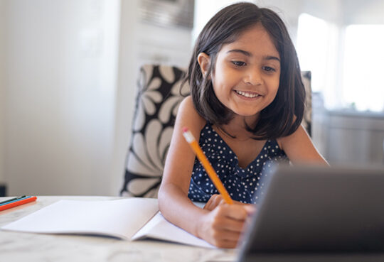 elementary age girl looking at a tablet smiles while writing on open notebook