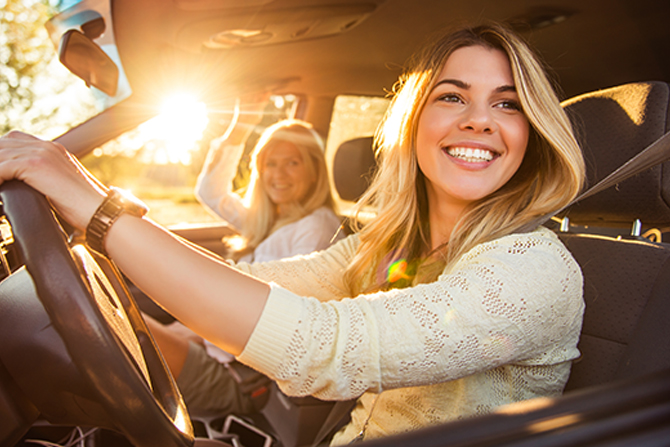 Two women in a vehicle during sunset, with the driver smiling and the passenger holding the car's interior grip handle