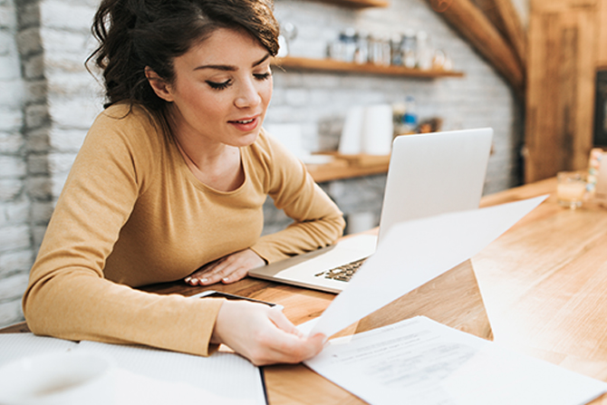 woman at kitchen counter looking at laptop and papers