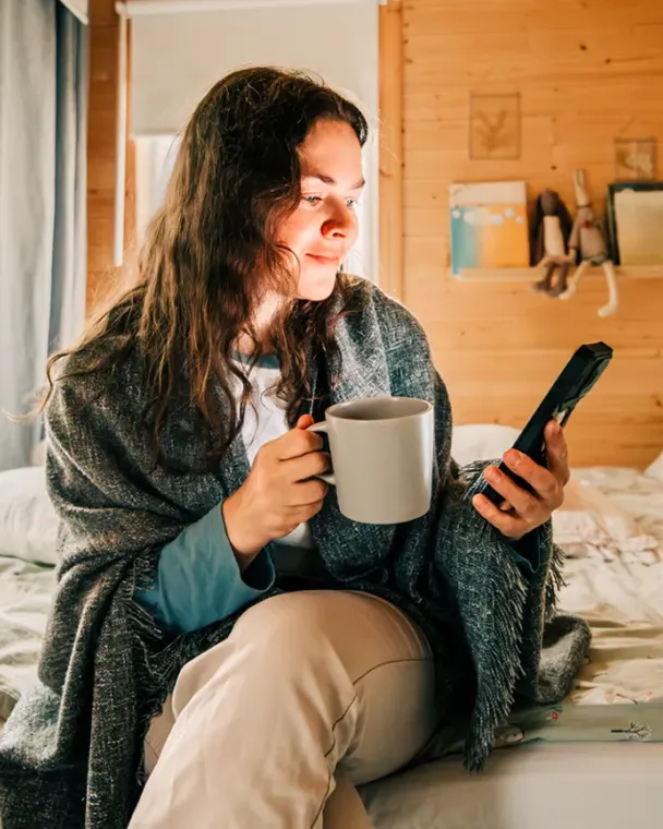 Woman sitting on bed looking at her phone and holding a coffee mug